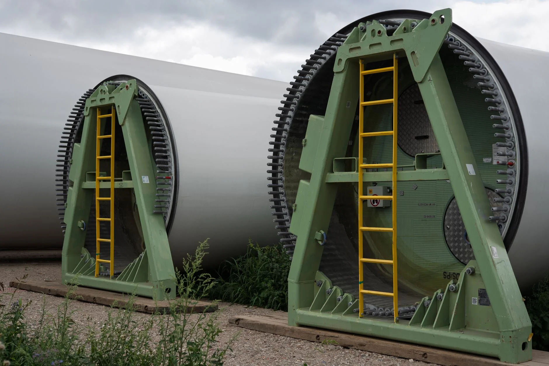 Wind turbine blade roots secured in steel transport frames at a staging yard, bolted interface rings marking where one supplier's scope ends